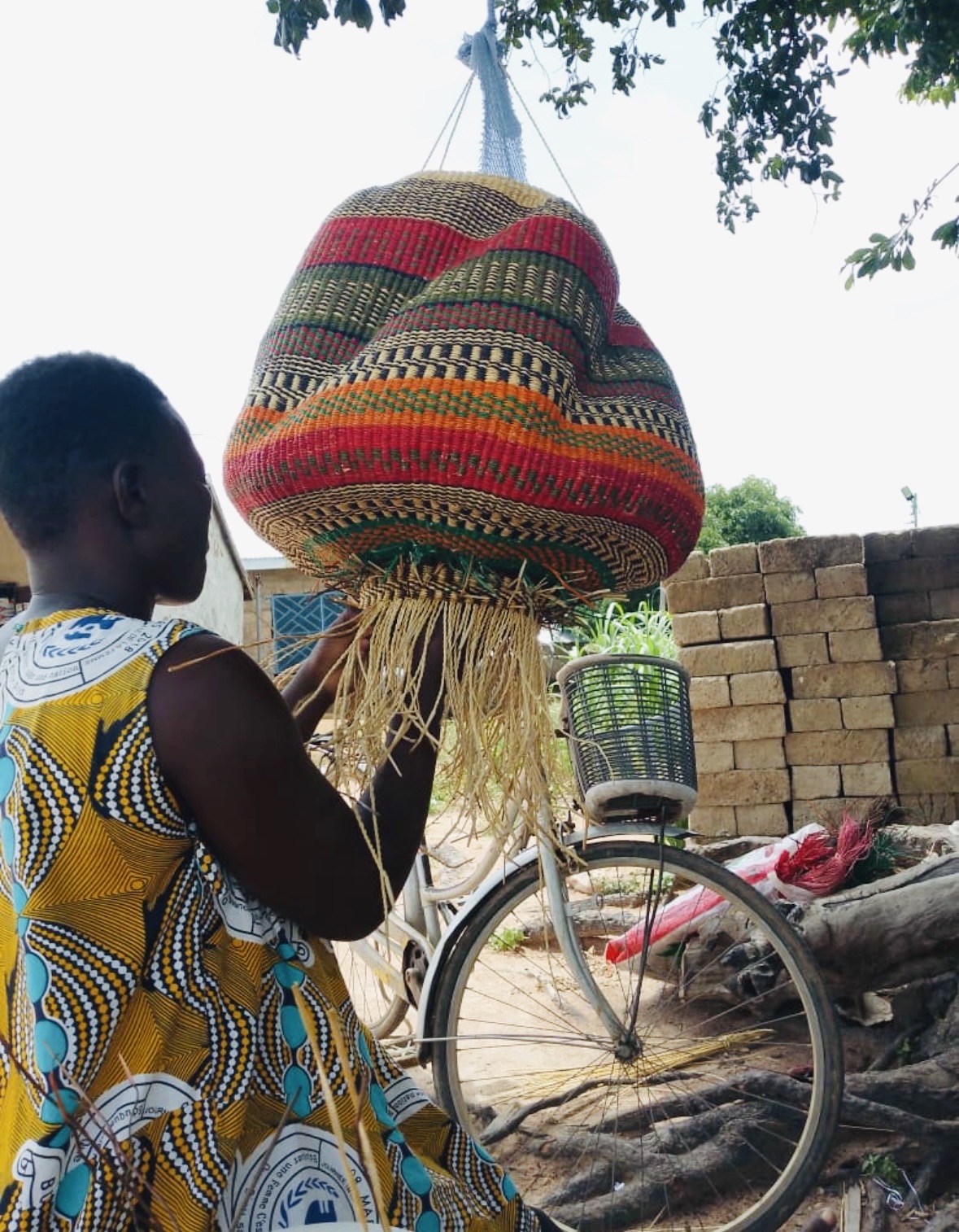 A maker steadying a large multi-coloured handwoven basket hung in a net outside in the village